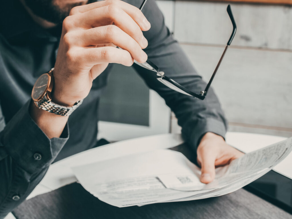 Mann sitzt Zuhause am Tisch, hält Papiere in einer Hand und hat in der anderen seine Brille: Titelbild zu Social Engineering bei Remote Work im Homeoffice